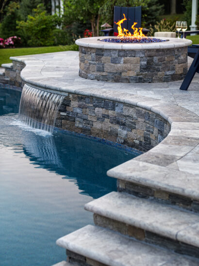 Travertine pool deck with water feature into pool and elevated fire pit deck. Dex by Terra residential work in Norfolk, MA.