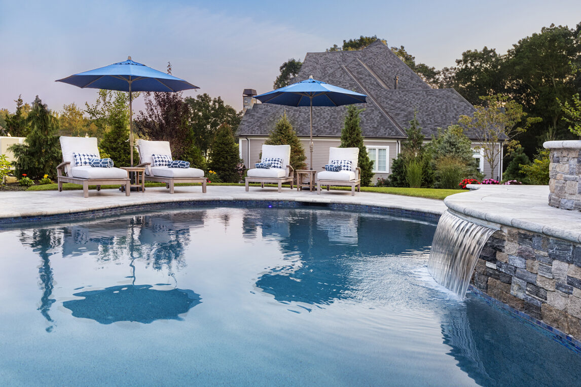 Pool chairs and water feature on a travertine pool deck built by Dex by Terra in Norfolk, Massachusetts.