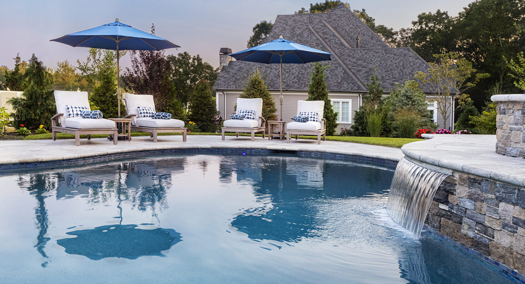 Pool chairs and water feature on a travertine pool deck built by Dex by Terra in Norfolk, Massachusetts.