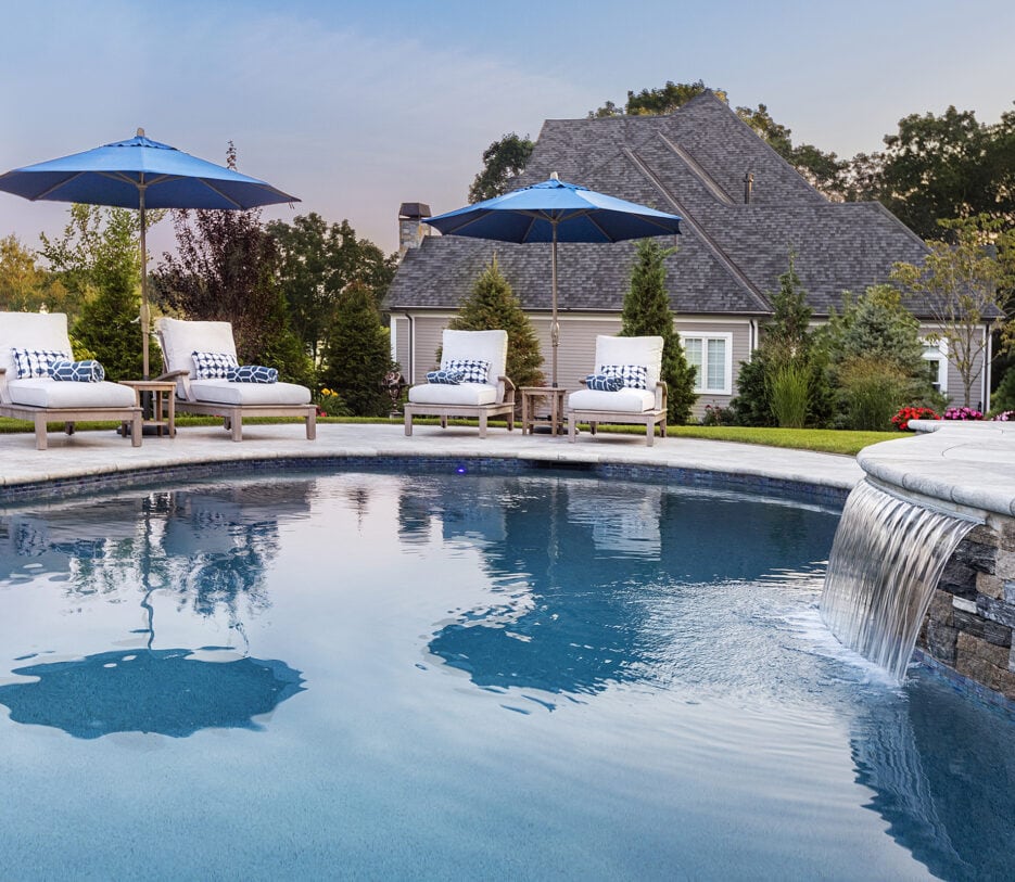 Pool chairs and water feature on a travertine pool deck built by Dex by Terra in Norfolk, Massachusetts.