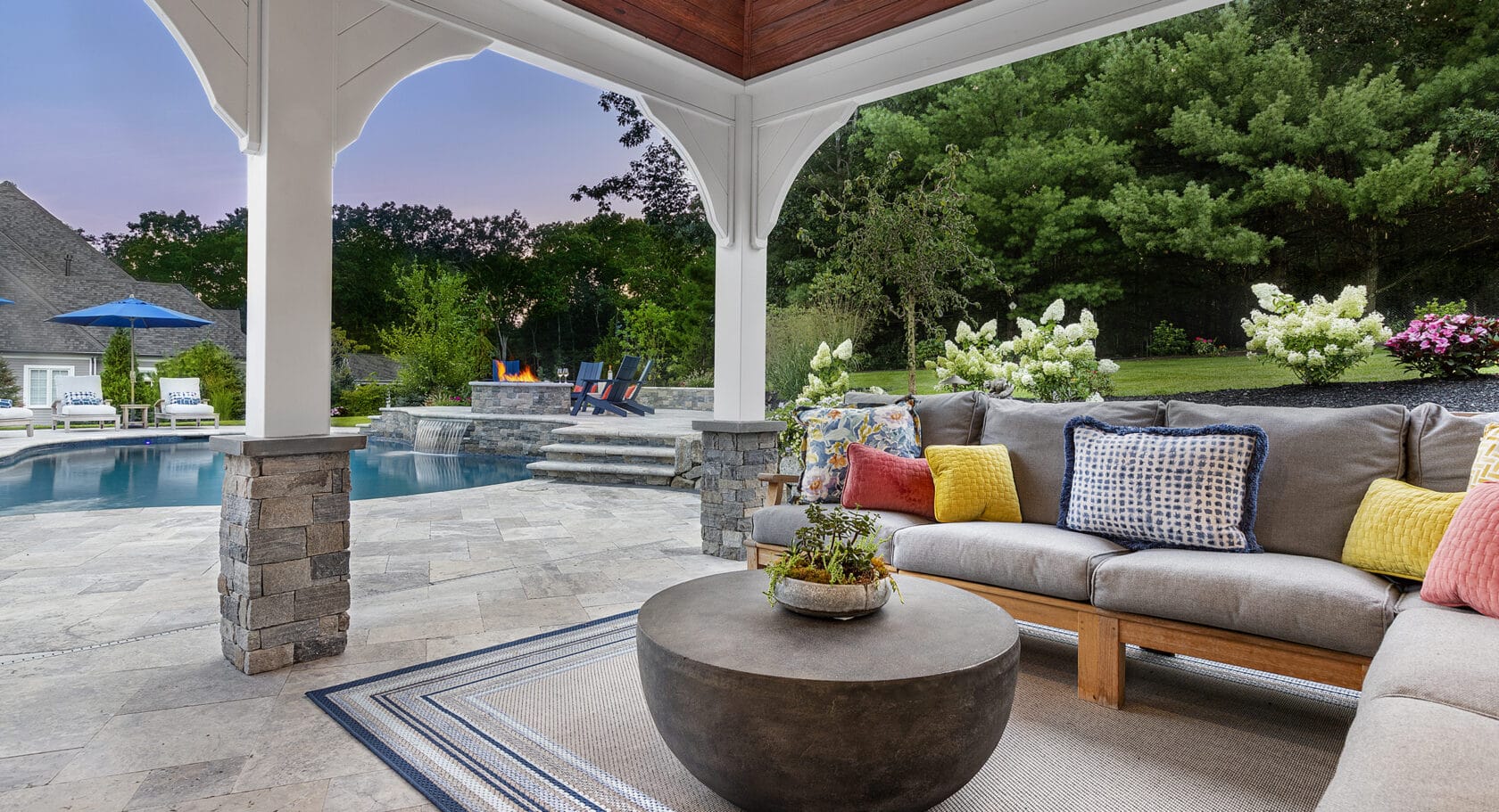 Pergola with couch and patio decor with pool in background. Landscape design-build by Dex by Terra in Norfolk, Massachusetts.