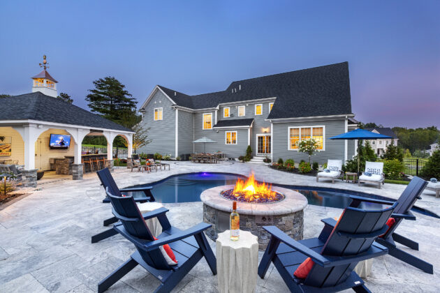 Fire pit with wine. Pool, patio, & pergola with outdoor kitchen in the background. Dex by Terra Hardscape in Norfolk, MA.
