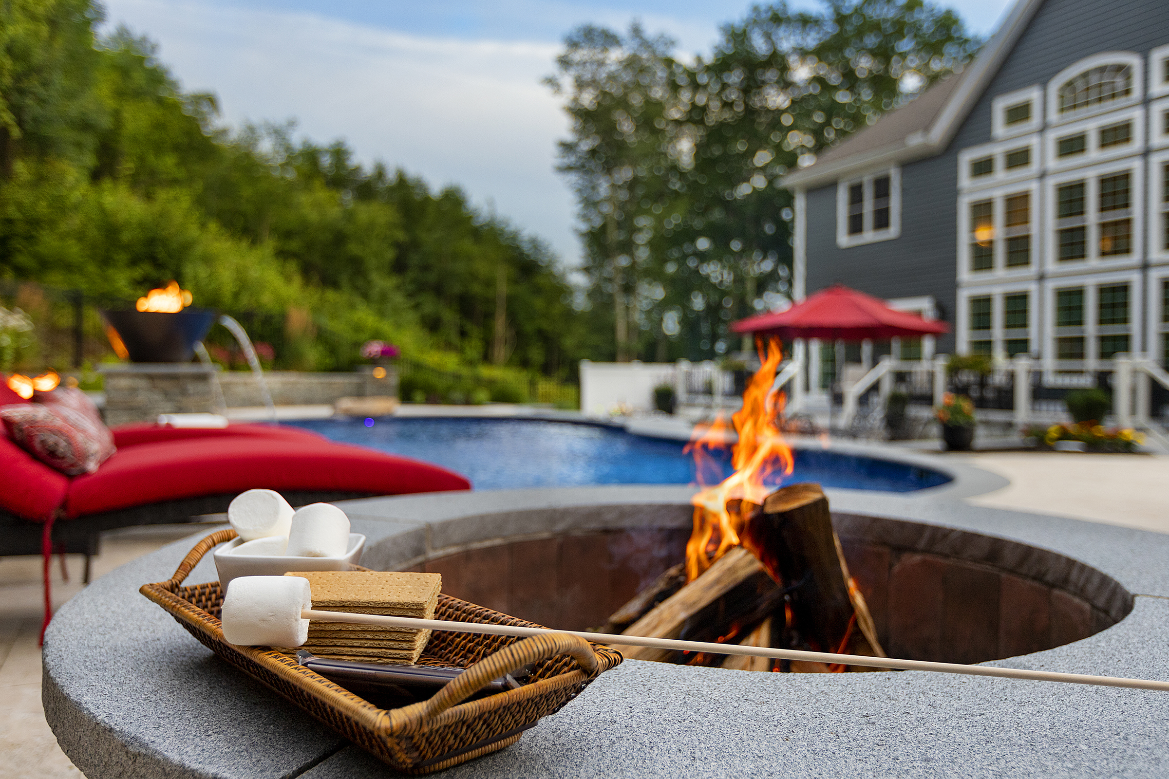 S'more kit by a wood-burning fire pit. Pool, fire bowl, and home in background. Dex by Terra Hardscaping in Sterling, MA.