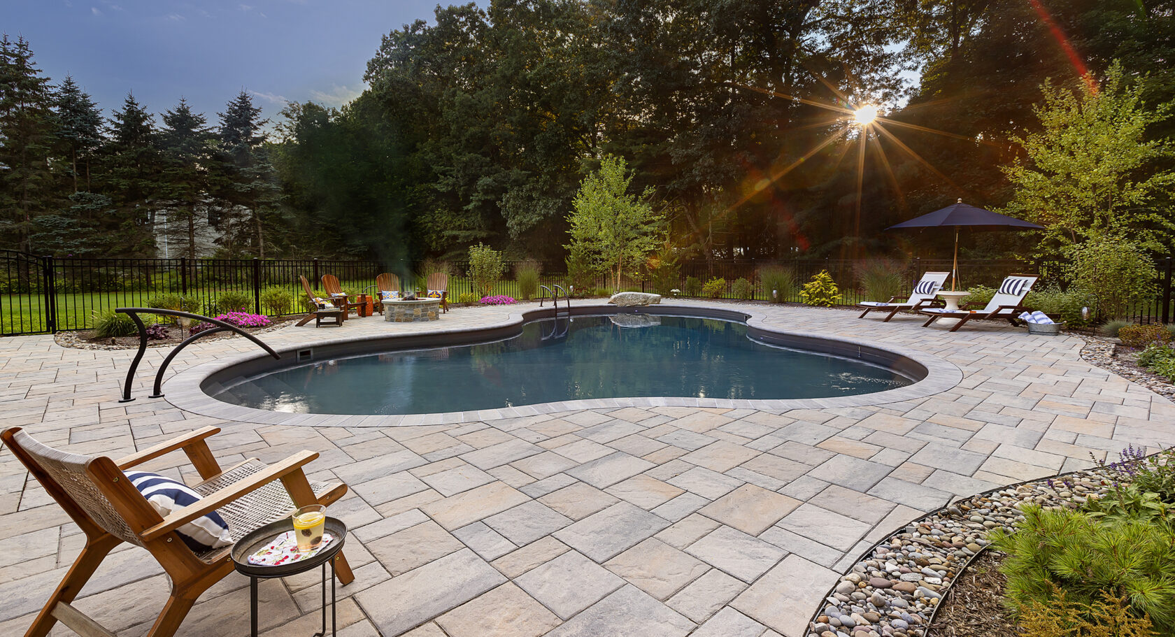 Pool chair overlooks inground pool with sunset through the trees. Paver pool deck design-build by Dex by Terra.