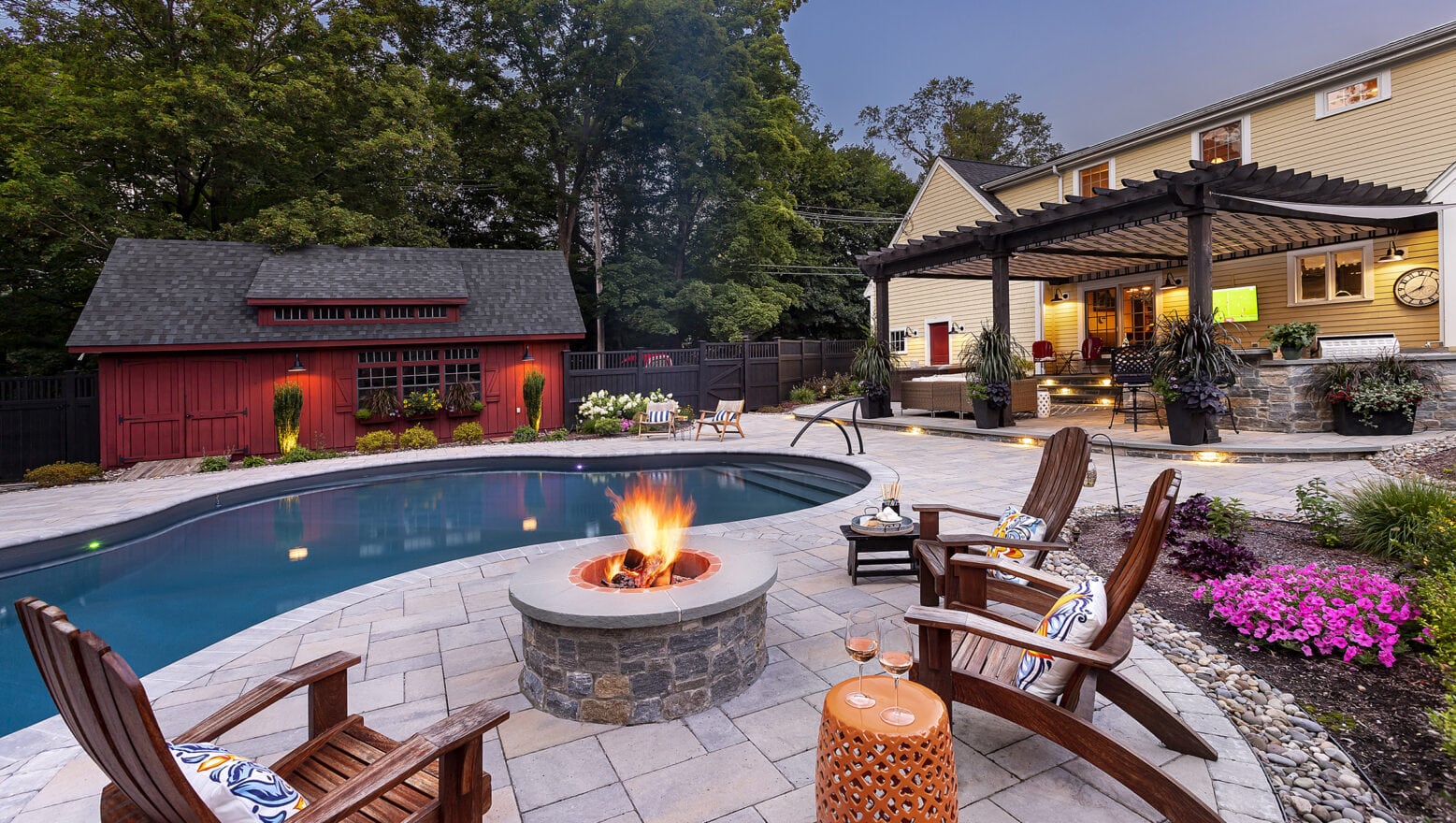 Poolside fire pit with barn and outdoor kitchen in background. Dex by Terra Landscape Design & Build Project in Stow, MA.