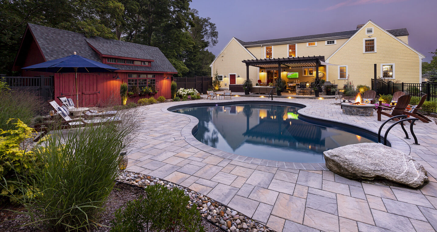 Unilock paver pool deck. Red barn & outdoor kitchen in the background. Dex by Terra Hardscape & Landscape in Stow, MA.