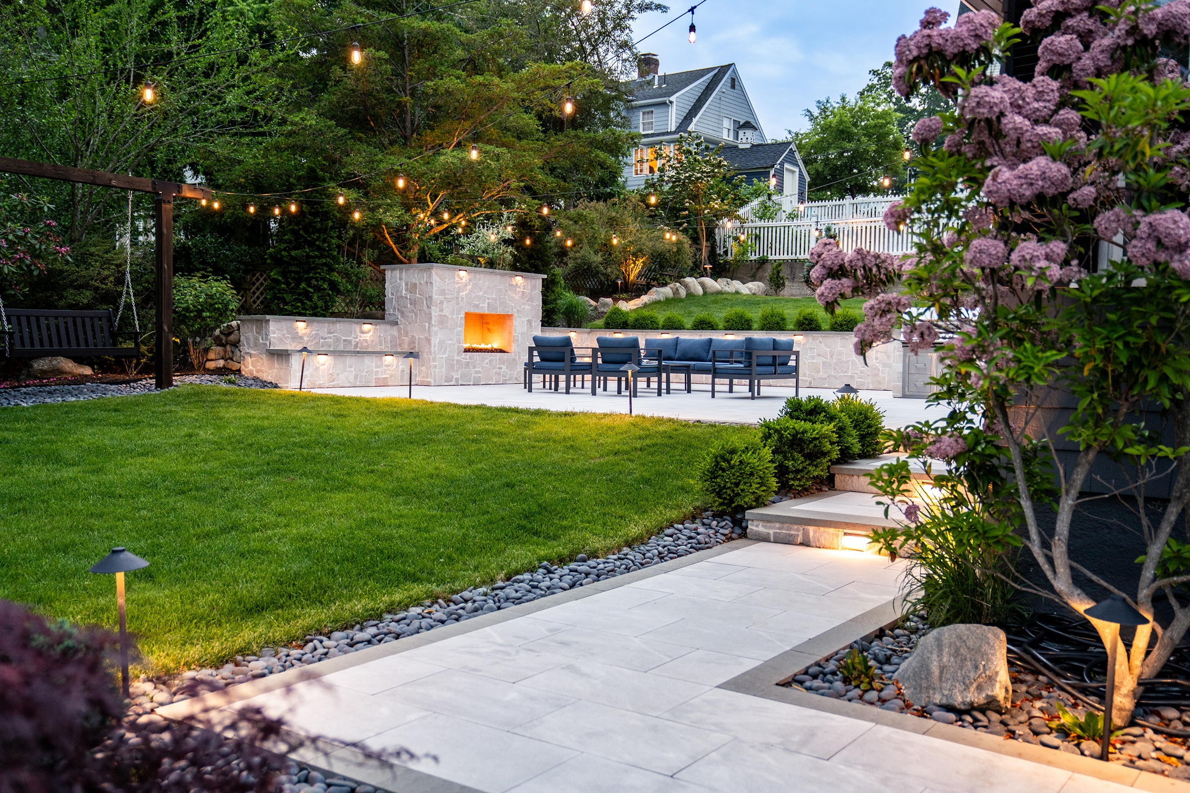 A natural stone paver walkway leads to a backyard patio with outdoor gas fireplace, patio furniture, and outdoor kitchen in Needham, MA.