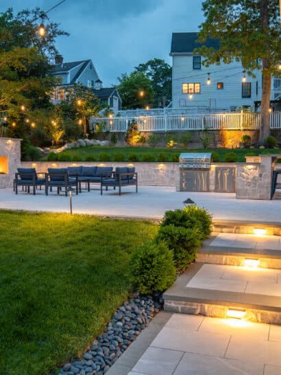 A natural stone paver walkway leads to a backyard patio with outdoor gas fireplace, and outdoor kitchen, illuminated by string lights and landscape lighting in Needham, MA.
