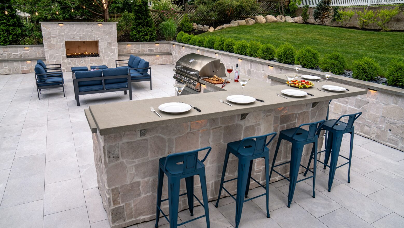 Outdoor kitchen with barstools overlooking patio furniture and an outdoor gas fireplace in Needham, MA.