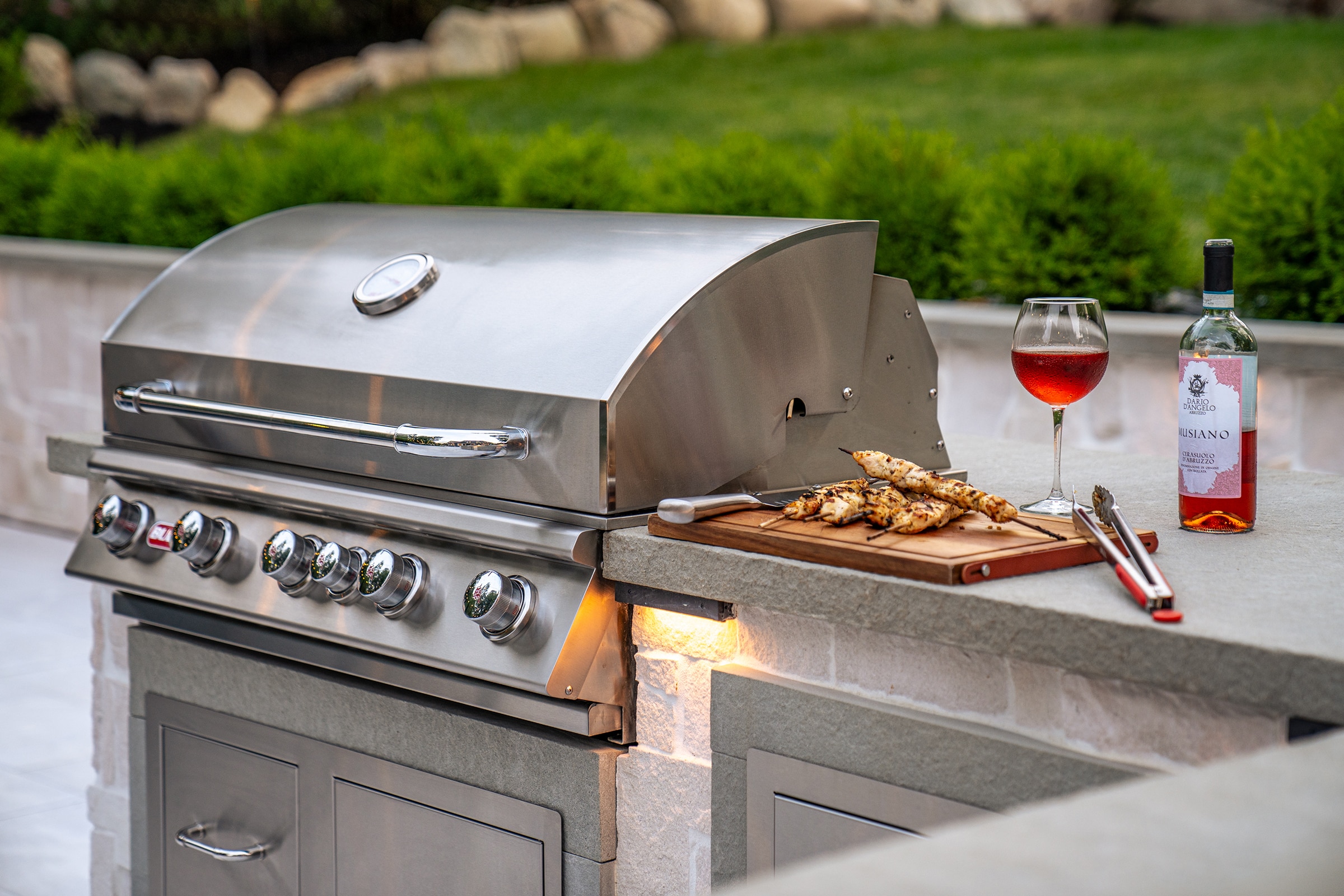 An outdoor kitchen with stainless steel grill and freshly prepared grilled chicken with red wine.