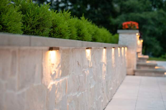 Stonewall with shrubs and low voltage outdoor landscape lighting in a Needham, MA backyard patio.