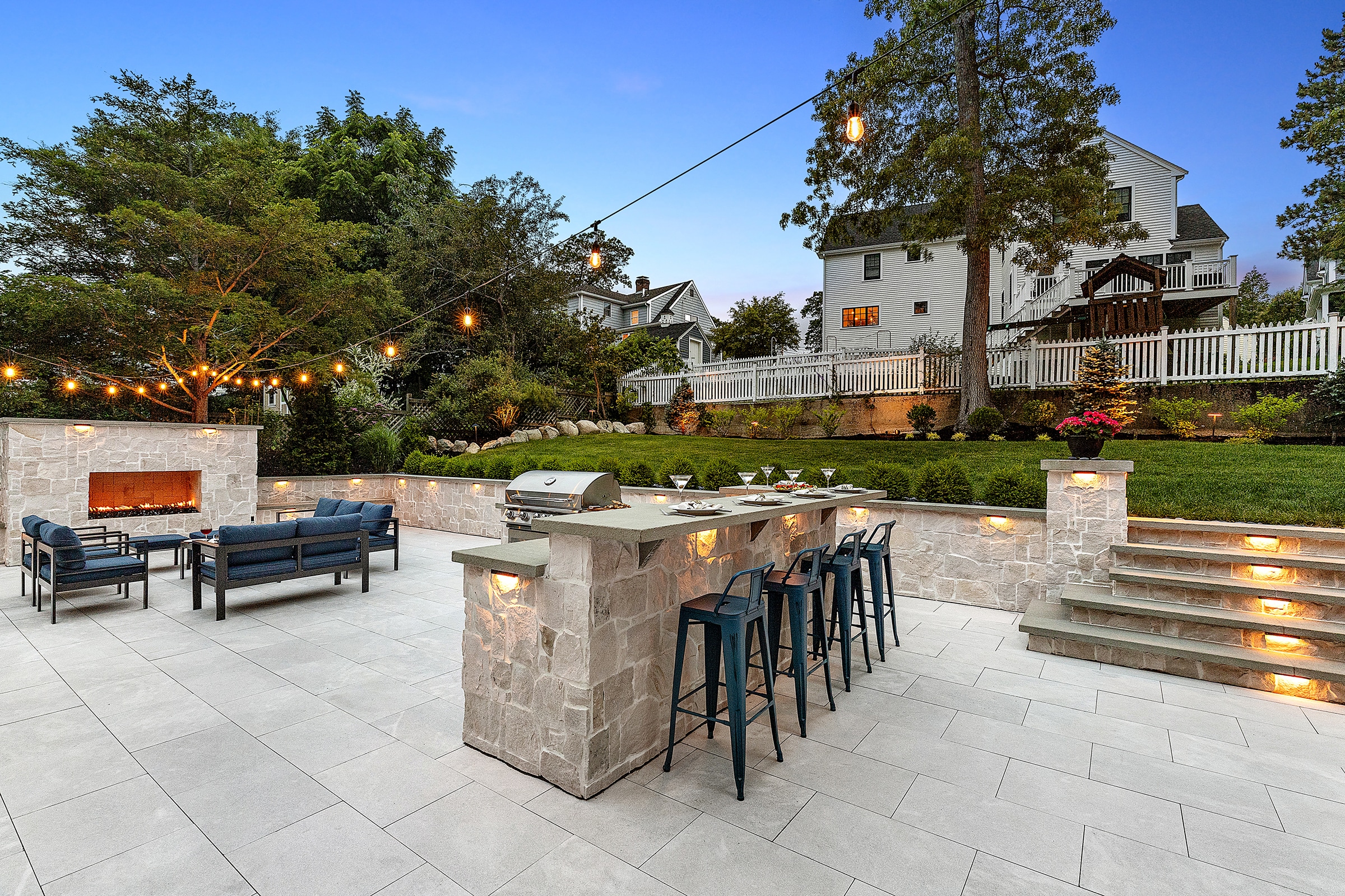 Backyard patio in Needham, Massachusetts with gas fireplace, outdoor kitchen, and stone stairs with outdoor lighting.