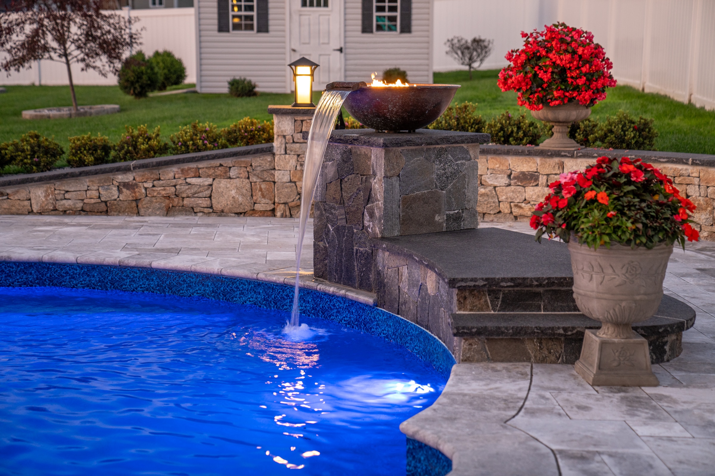 A pool deck with water and fire bowls mounted on a stone veneer column with natural stone caps. Dex by Terra hardscaping project in Grafton, MA.